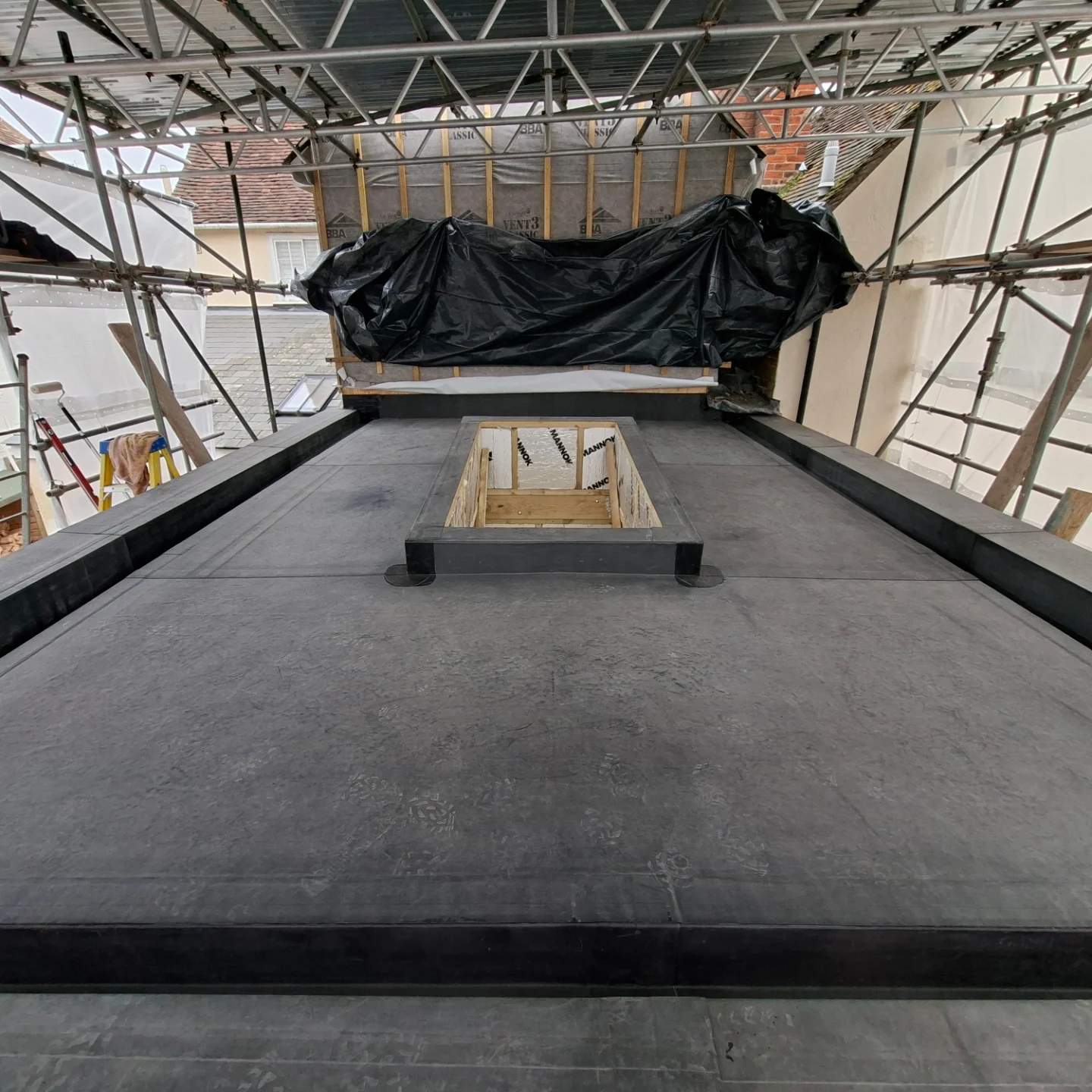 A finished black rubber flat roof with a rooflight upstand, under scaffold